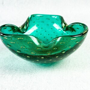 May include: A teal-colored glass ashtray with a unique shape. The ashtray has a wide, shallow bowl with a slightly raised rim and a textured surface with small, gold-colored inclusions. The overall design is elegant and decorative.
