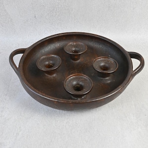 May include: A round, dark brown ceramic candle holder featuring two side handles and four integrated, circular candle cups arranged in a central cluster. The surface has a mottled, earthy texture with subtle variations in the deep brown glaze.