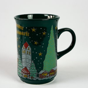 May include: A dark green ceramic mug with a festive winter scene. The mug features a church, Christmas tree, and small houses, all decorated with gold stars and snow. The mug has a black handle and the words "Wunder Weihnachtsmarkt" in gold.