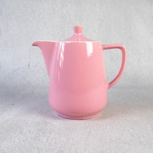 Könnte beinhalten: Eine rosa Keramik-Teekanne mit Deckel und Griff. Die Teekanne hat einen gebogenen Ausguss und einen abgerundeten Körper. Der Griff hat eine einfache Schlaufenform. Die Teekanne hat eine einfarbige, glänzende rosa Farbe.