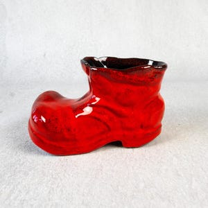 Könnte beinhalten: Ein roter Keramik-Pflanztopf in Form eines Holzschuhs. Der Stiefel hat eine glänzende Oberfläche mit dunkelbraunen Akzenten am Rand. Dieser dekorative Artikel eignet sich für kleine Pflanzen oder als einzigartiges Wohnaccessoire.