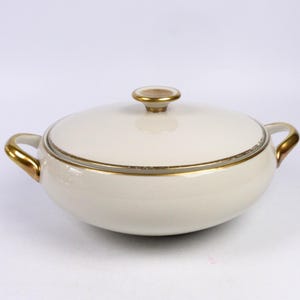 Può includere: Un piatto da portata in porcellana bianca con un bordo e manici dorati. Il piatto ha un coperchio con un pomo dorato.