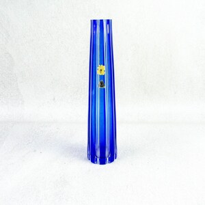 Op de afbeelding: Een hoge, blauwe glazen vaas met verticale ribbels. De vaas heeft een slanke, taps toelopende vorm en een kleine, ronde basis. Er is een gouden en zwarte sticker op de voorkant bevestigd. De vaas is waarschijnlijk voor decoratieve doeleinden.
