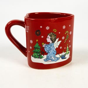 May include: A red ceramic mug with a curved handle, featuring a Christmas angel illustration. The angel is depicted in a blue robe, decorating a Christmas tree. Snowflakes and candy canes decorate the mug's exterior.