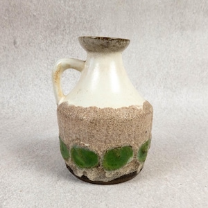 May include: A ceramic pitcher featuring a smooth off-white upper section and a textured, sandy-beige lower body. The base is decorated with a horizontal row of circular, glossy green glazed spots. It has a single handle and a flared rim.