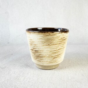 Può includere: Una piccola tazza in ceramica beige con un motivo strutturato simile alla corteccia. Il bordo è di colore marrone scuro e la tazza ha una forma leggermente svasata. La tazza viene probabilmente utilizzata per bere bevande.