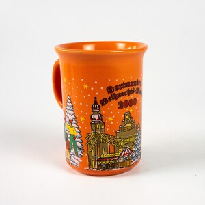 May include: Orange ceramic mug with a handle, depicting a winter scene with a church, trees, and figures. The mug features the text "Dortmunder Weihnachtsmarkt 2000" and star accents.