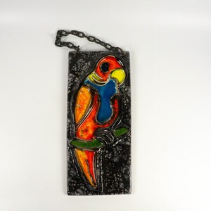 May include: A ceramic wall hanging depicting a colourful parrot perched on a branch. The parrot is painted in shades of orange, red, blue, yellow, and green. The background is black with a speckled texture.