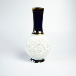 May include: A white porcelain vase with a blue and gold rim and a raised white floral design. The vase has a round body and a long, narrow neck.