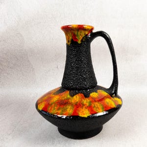 May include: A black ceramic vase with a textured surface and a handle. The vase features a vibrant design with orange, yellow, and red hues on the base and rim. The vase is approximately 10 inches tall.