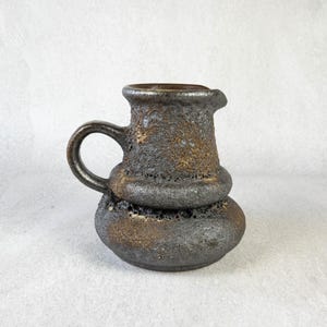 May include: A small, gray and brown ceramic pitcher with a textured surface. The pitcher has a handle and a rounded body.