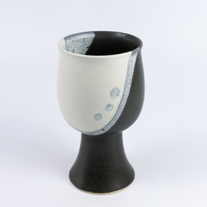 Può includere: Un calice in ceramica bianco e nero con una base larga e una ciotola arrotondata. La ciotola ha una smaltatura bianca con un vortice nero e macchie blu.