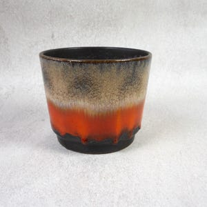 May include: A ceramic planter with a striking colour gradient. The top is black, transitioning to tan, white, and vibrant orange at the base. The planter has a textured surface and a rounded shape.