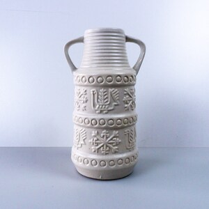 May include: A white ceramic vase with two handles and a geometric pattern. The pattern includes snowflakes, birds, and circles.