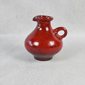 May include: A small, bulbous ceramic pitcher with a glossy, deep red glaze. The vessel features a rounded body, a short neck with a small pouring spout, and a single curved handle attached to the side. The finish shows subtle tonal variations.