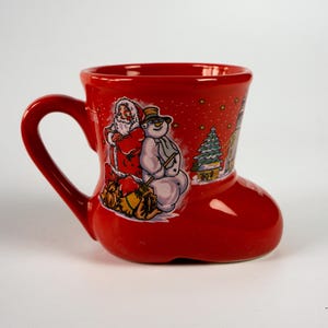 May include: A red ceramic boot-shaped mug with a handle. The mug features a festive illustration of Santa Claus, a snowman, and a winter scene. The mug is designed for holiday use.