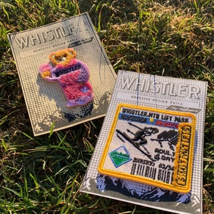 Pode incluir: Duas aplicações adesivas de design na relva. Uma aplicação apresenta um urso a usar um traje rosa e roxo com a palavra "Whistler". A outra aplicação é um passe de teleférico com borda amarela com o texto "Whistler Mtn Lift Pass". Ambas as aplicações têm a palavra "Whistler".