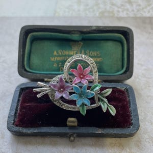May include: Vintage heart-shaped brooch with three colorful enamel flowers in pink, blue, and purple. The brooch is set with small clear stones and is displayed in a black velvet-lined box.