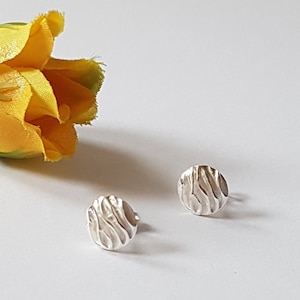 May include: A pair of small, silver-colored stud earrings with a textured, wave-like pattern. The earrings are round and sit on a white surface, with a yellow flower in the background.
