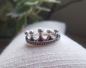 Könnte beinhalten: Ein silberner Kronenring mit einer strukturierten Band und fünf Spitzen. Der Ring liegt auf einer weißen Stoffoberfläche.