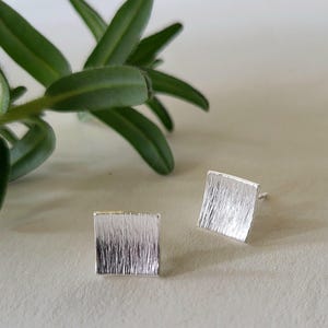 May include: A pair of square silver stud earrings with a textured, brushed metal finish. The earrings are displayed on a light-colored surface, with green leaves in the background. The earrings are approximately 0.5 inches square.