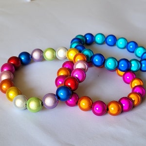 Könnte beinhalten: Drei bunte Armbänder aus runden Perlen. Die Armbänder sind übereinander gelegt. Das obere Armband ist blau, das mittlere Armband ist bunt gemischt und das untere Armband ist bunt gemischt.