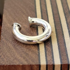 Könnte beinhalten: Ein silberfarbener, gebogener Ohrring mit einem abgerundeten, röhrenförmigen Design. Der Ohrring hat eine glatte, polierte Oberfläche und eine kleine, kreisförmige Öffnung an jedem Ende. Der Ohrring liegt auf einer Holzoberfläche mit abwechselnd hellen und dunklen braunen Streifen.