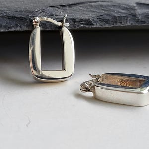 May include: A pair of silver-colored, rectangular hoop earrings. One earring is upright, showing the open rectangular shape, while the other lies on its side. The earrings have a polished finish and a simple, modern design.