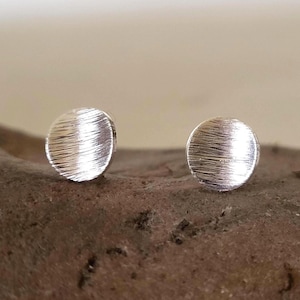 Könnte beinhalten: Zwei kleine, runde Ohrstecker aus Silber. Jeder Ohrring hat eine strukturierte, gebürstete Metalloberfläche. Die Ohrringe werden auf einer rauen, braunen Oberfläche mit einem weichen, neutralen Hintergrund präsentiert.