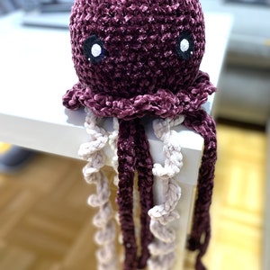 May include: A crocheted plush jellyfish toy with a purple body and white eyes. The jellyfish has long, curly tentacles in shades of purple and white.