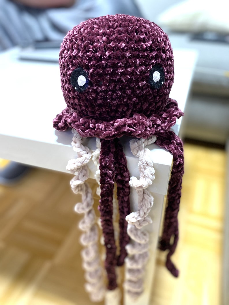 May include: A crocheted plush jellyfish toy with a purple body and white eyes. The jellyfish has long, curly tentacles in shades of purple and white.