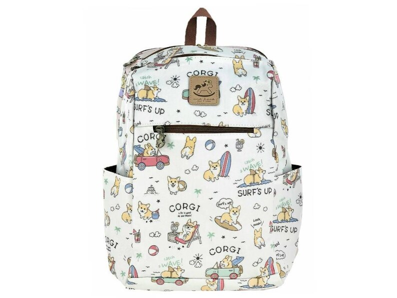 May include: White canvas backpack featuring a colourful pattern of corgis surfing, driving cars, and relaxing on the beach. The backpack has a brown leather handle and zipper pulls. The text "CORGI" and "SURF'S UP" is repeated throughout the pattern.