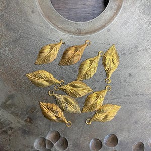 May include: A collection of gold-colored metal leaf charms with a loop on each leaf. The charms are arranged on a gray metal surface.