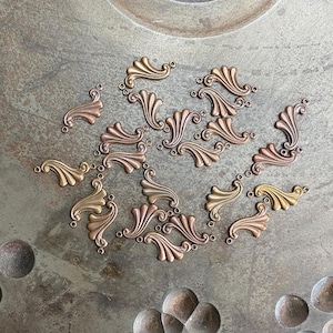 May include: A collection of small, decorative metal swirls in gold and copper tones arranged on a metal surface. The swirls have a delicate, ornate design and are likely used for crafting or jewelry making.