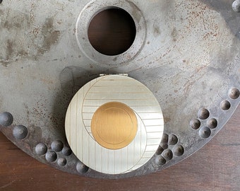 May include: A silver and gold compact mirror with a swirling design. The mirror is sitting on a large, round, metal disc with a textured surface.