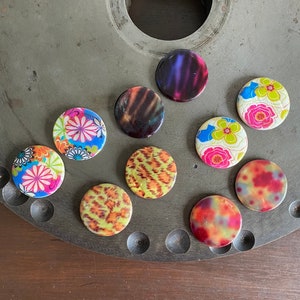May include: Nine colorful round buttons with various patterns, including floral, animal print, and abstract designs. The buttons are arranged on a gray metal surface.