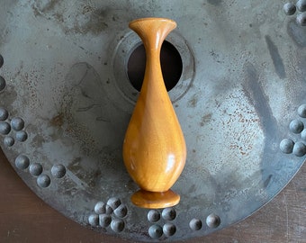 May include: A wooden vase with a light brown color and a rounded shape. The vase is sitting on a gray metal surface with a pattern of small circles.