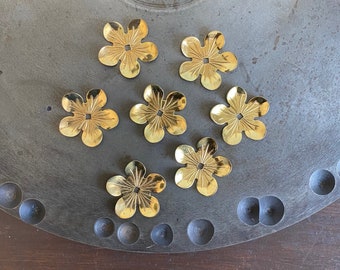Può includere: Sette componenti metallici dorati a forma di fiore con dettagliati disegni di petali sono disposti su una superficie metallica grigia e circolare. I fiori hanno un foro quadrato centrale e sono uniformemente distanziati.