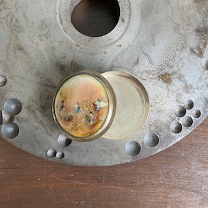 Puede incluir: Una pequeña caja redonda de latón con una tapa con bisagras. La tapa está decorada con una escena colorida de personas montando a caballo. La caja está sobre una superficie metálica.