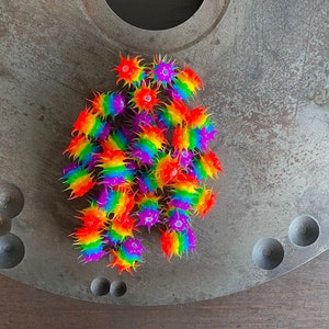May include: A collection of small, colorful, spiky toys arranged on a textured, gray metal surface. The toys display a rainbow of colors, including red, orange, yellow, green, blue, and purple. The metal surface has a large circular indentation.