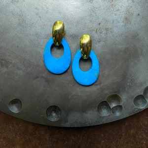 May include: A pair of blue and gold earrings with a circular design. The earrings are on a gray metal surface.