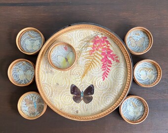 Könnte beinhalten: Ein Set aus acht Untersetzern mit Schmetterlings- und Farnmotiven. Die Untersetzer sind aus Holz und haben eine hellbraune Oberfläche. Die Untersetzer sind in einem Kreis um einen größeren Untersetzer mit einem Schmetterlings- und Farnmotiv angeordnet.