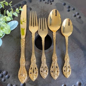 May include: A set of five gold-colored flatware pieces, including a knife, two forks, and two spoons. The handles feature an ornate floral design. The pieces are arranged on a dark, textured surface.
