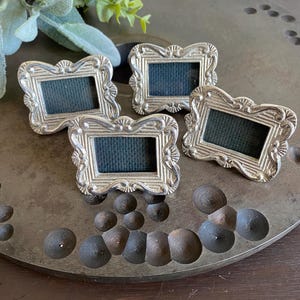 Puede incluir: Cuatro pequeños marcos de fotos plateados y ornamentados con inserciones azul oscuro están dispuestos sobre una bandeja decorativa de metal. Los marcos tienen un diseño detallado de estilo vintage. La bandeja tiene una superficie texturizada y está decorada con acentos oscuros y redondeados.