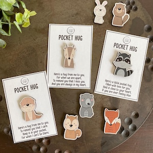 May include: Three white rectangular cards labeled POCKET HUG with wooden animal pins attached, including a deer, an otter, and a raccoon. Loose wooden pins of a rabbit, beaver, bear, squirrel, and fox are arranged around the cards on a dark tray.