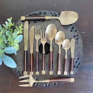 May include: A collection of vintage brass flatware with dark wood handles. The set includes knives, forks, spoons, and a serving spoon. The utensils are arranged on a dark, textured surface, showcasing their classic design.