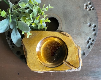 May include: A natural stone ashtray with a circular depression and a cigarette rest. The ashtray is a warm, golden-brown colour, with a polished interior. A small bundle of green foliage rests nearby.