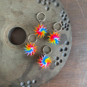 May include: Five colorful, spiky starburst keychains. Each keychain features a vibrant mix of rainbow colors, including red, orange, yellow, green, blue, and purple. The keychains are attached to silver-colored key rings.
