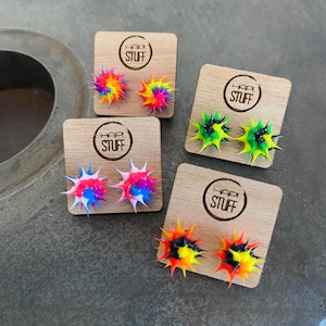 May include: Four pairs of colorful silicone stud earrings on wooden display cards. Each pair features a different color combination, including rainbow, green and black, pink and blue, and orange and black. The cards have the text "Hap Stuff" printed on them.