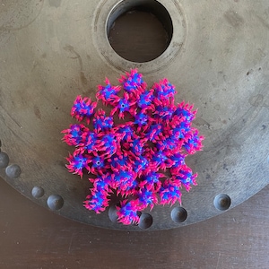 Op de afbeelding: Een groep kleine, stekelige, blauwe en roze rubberen ballen. De ballen zijn gerangschikt in een cluster op een grijs metalen oppervlak.